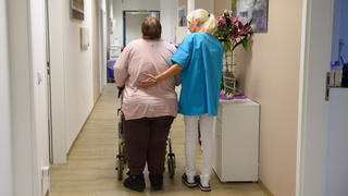 Pflegerin und Patientin mit Rollator bei einem Spaziergang über den Flur der Pflegeeinrichtung.