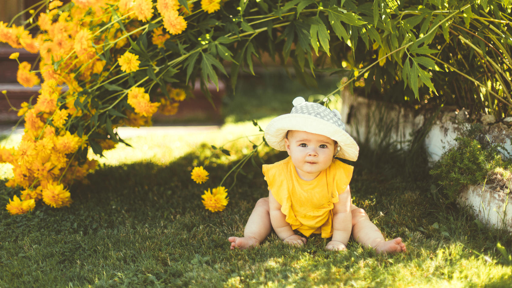 Baby mit gelbem Kleid und Sonnenhut sitzt im Sommer im Garten