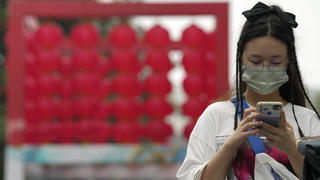 14.08.2022, China, Beijing: Eine Frau, die eine Mund-Nasen-Schutz trägt, überprüft in der Nähe eines Einkaufszentrums ihr Telefon. China hat die höchsten Corona-Infektionszahlen seit über drei Monaten gemeldet. Foto: Ng Han Guan/AP/dpa +++ dpa-Bildfunk +++