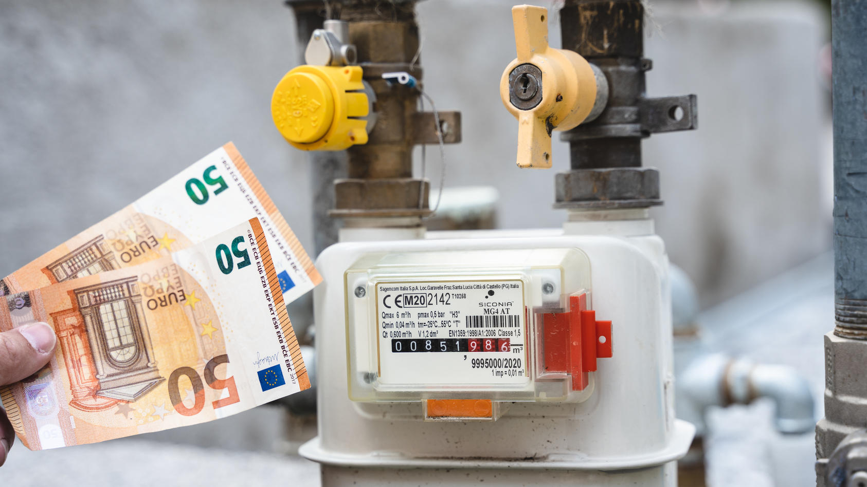 Riva, Trentino, Italy - 15 August 2022: Gas meter from a residential building outdoors. Male hand holding 50 euro bills in front of gas meter and pipe, symbolic image rising cost of electricity, gas and energy *** Gaszähler von einem Wohnhaus im Frei