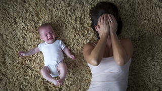 Eine Mutter liegt mit einem Baby auf dem Boden. Das Kind weint und die Mutter hält sich die Hände vor dem Kopf.