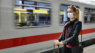 Frau mit Mundschutz und Koffer steht vor Zug