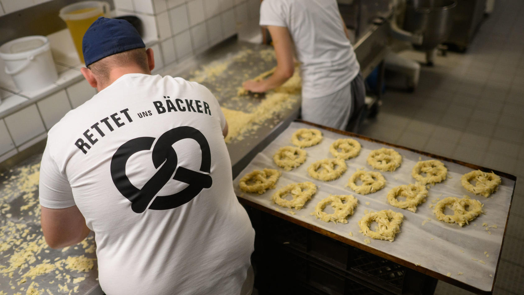 Ein Bäcker und eine Auszubildende stehen am frühen Morgen mit einem T-Shirt ·Rettet uns Bäcker· in der Backstube