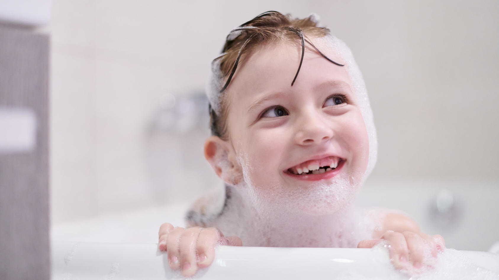 Kind schaut lachend und voller Schaum über den Rand der Badewanne