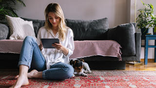Eine Frau sitzt mit ihrem Hund an der Seite auf dem Boden und schaut auf ein Tablet.
