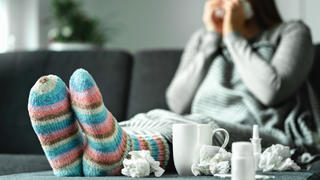 Eine junge Frau sitzt krank auf ihrem Sofa, um sie herum sind unter anderem ganz viele Taschentücher verteilt.