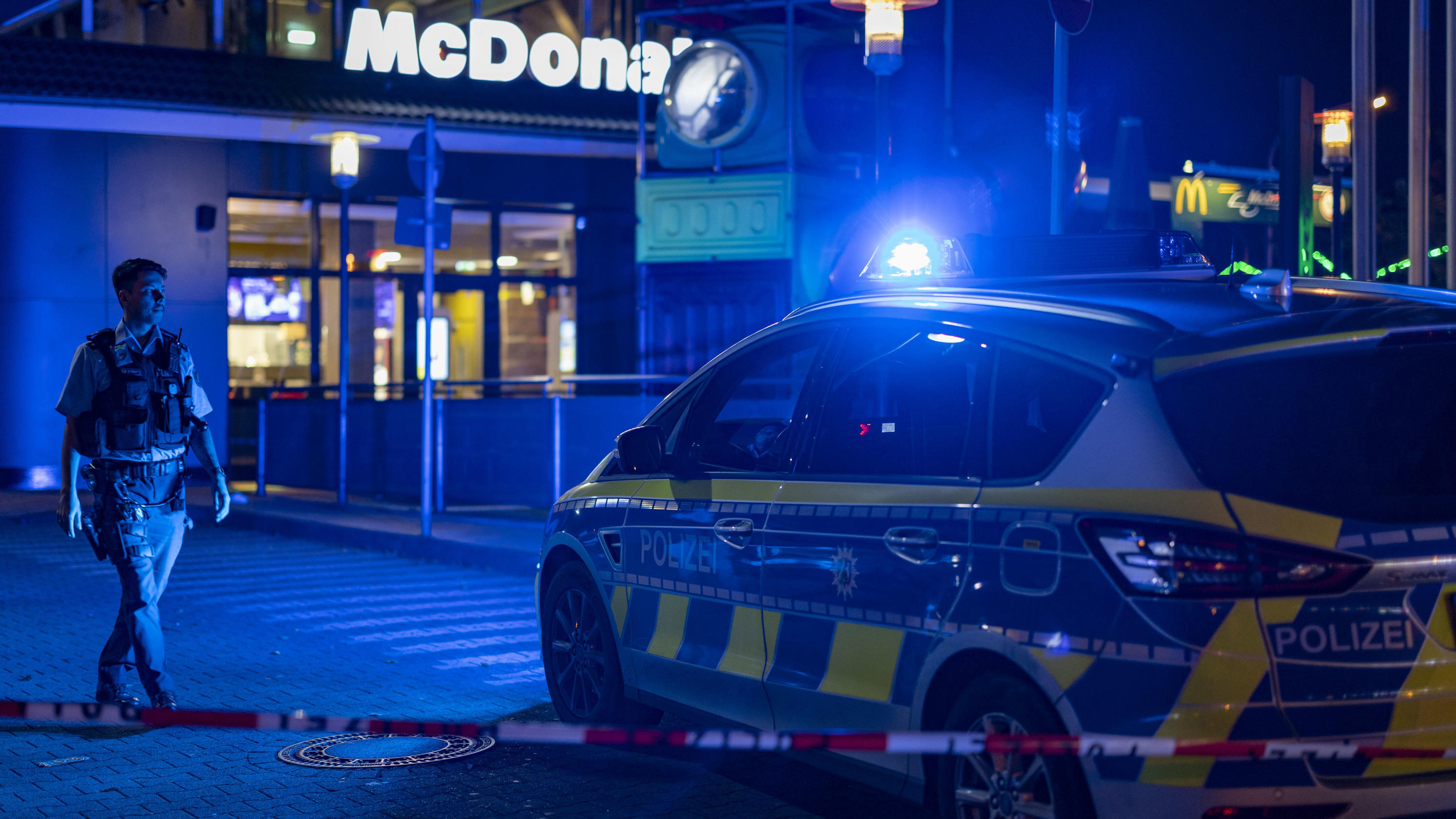 29.10.2022, Nordrhein-Westfalen, Oberhausen: Ein Polizist geht vor einem Fast-Food-Restaurant im Blaulicht, nachdem im Außenbereich Schüsse gefallen waren. Drei Menschen wurden verletzt. Der Täter sei auf der Flucht, teilte ein Polizeisprecher mit. D