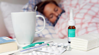 Frau liegt krank im Bett, vor ihr Medikamente auf dem Nachttisch.