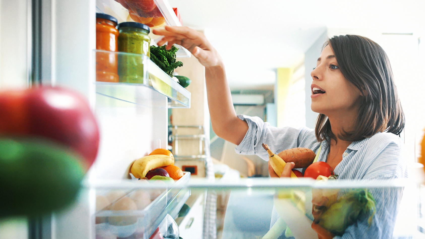 Una donna si trova davanti a un frigorifero e porta tra le mani o tra le braccia vari generi alimentari.