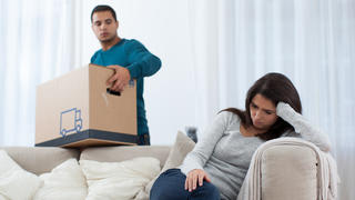 Mann packt nach Trennung Kartons, Frau sitzt traurig auf dem Sofa