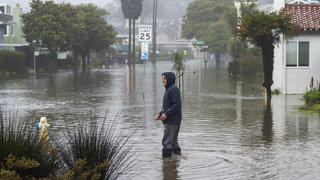09.01.2023, USA, Aptos: Ein Mann watet durch eine überschwemmte Straße in der Nähe von Rio Del Mar. Foto: Nic Coury/FR171100 AP/dpa +++ dpa-Bildfunk +++