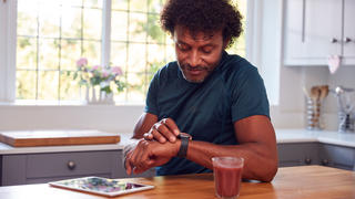 Mann guckt auf seine Smartwatch und sitzt mit Tablet in der Küche.