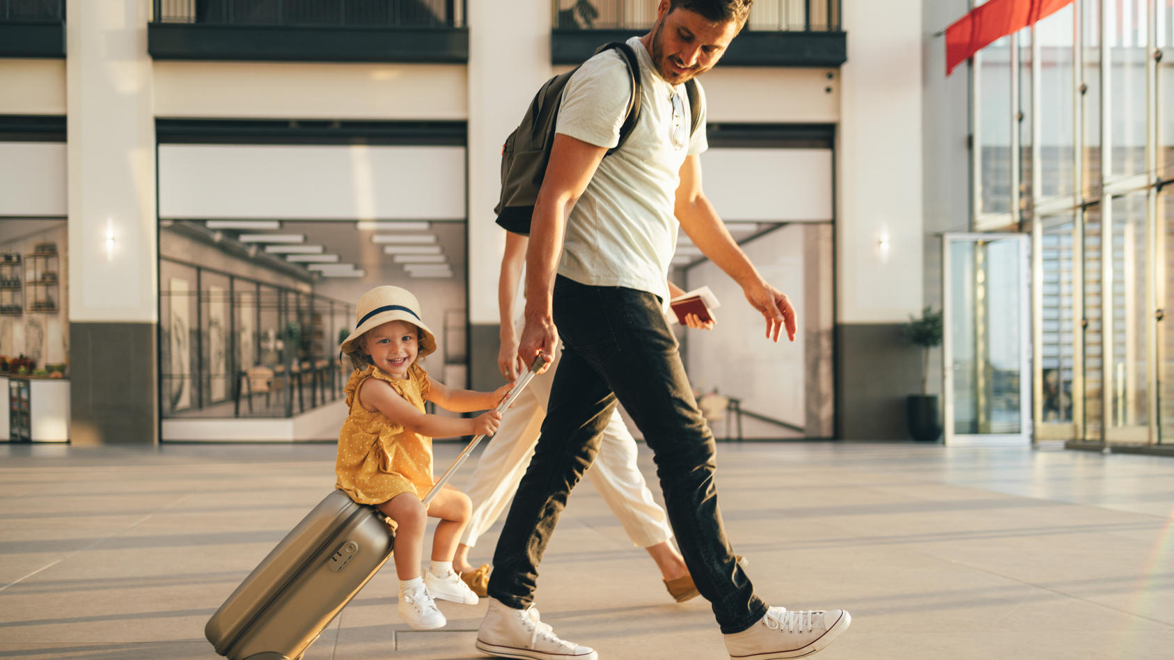 Ein junger Vater rollt seinen Handgepäckkoffer durch den Flughafen. Seine kleine Tochter sitzt auf dem Koffer.