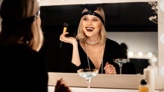 Eine blonde Frau sitzt in einem 20er-Jahr Outfit vor einem Spiegel und trägt Make-up auf.