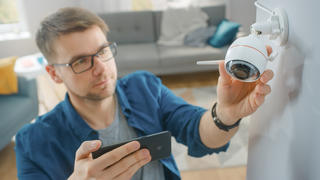 Junger Mann hängt eine Überwachungskamera in seinem Wohnzimmer auf. Leichte Installation durch die passende App.