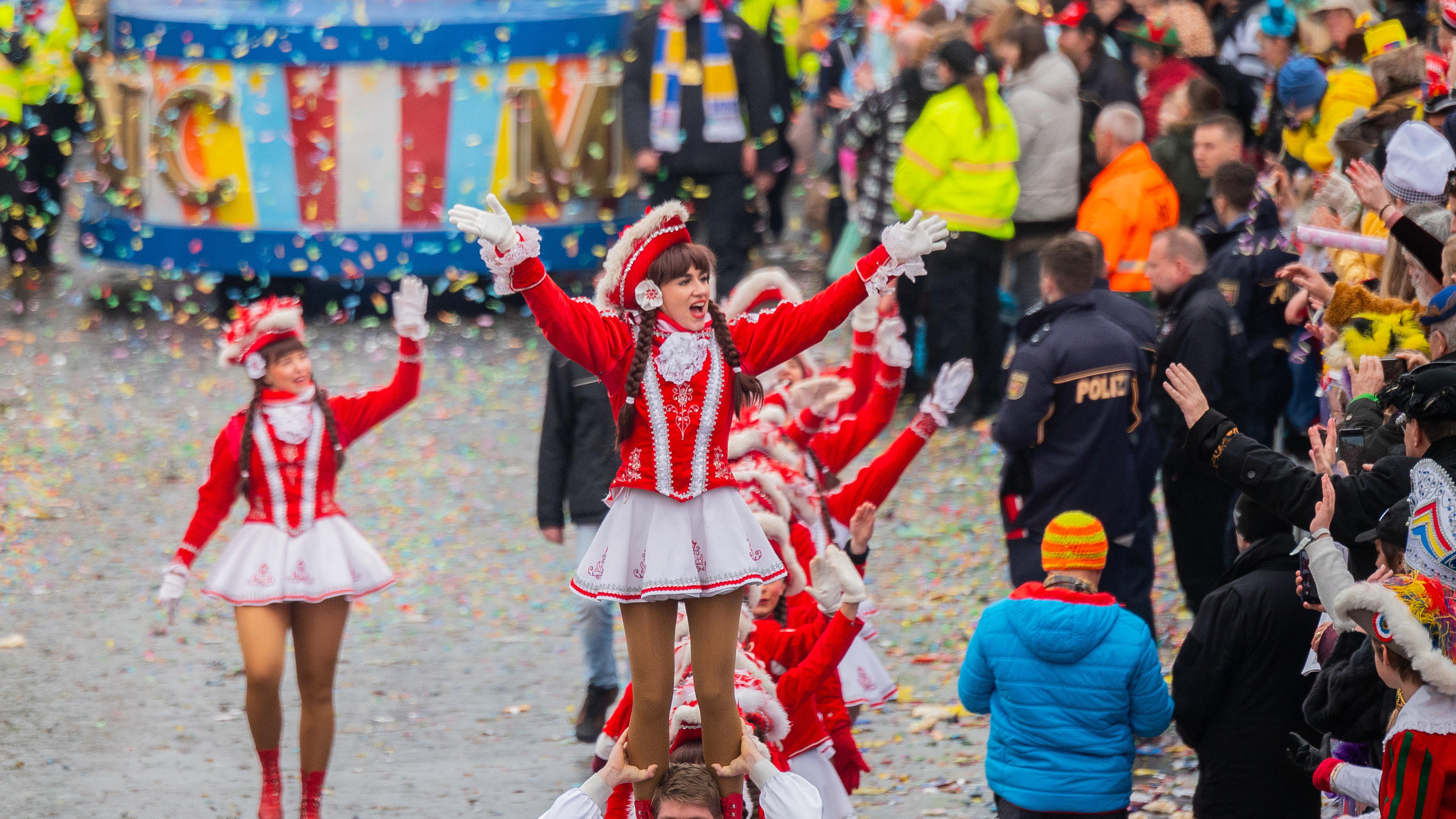 ARCHIV - 24.02.2020, Rheinland-Pfalz, Mainz: Ein Tanzmariechen des Mainzer Narren-Clubs (MNC) wird auf den Schultern getragen. Polizisten sichern den Rosenmontagszug, der zuletzt 2020 stattfand. In diesem Jahr bereiten Auflagen für Sicherheitsvorkehr