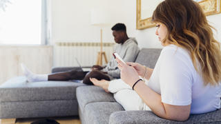 Frau auf Sofa schaut auf ihr Handy, Mann im Hintergrund ist am Laptop