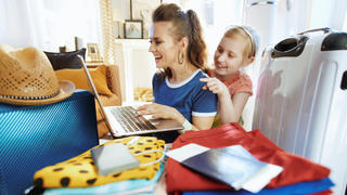 Eine lachende Mutter und ein Kind sitzen zuhause vor einem Laptop und neben vielen Urlaubsgegenständen und einem Reisekoffer.