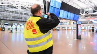 17.02.2023, Hamburg: Ein Flughafen-Mitarbeiter steht mit Streikweste im sonst menschenleeren Terminal 1 und fotografiert die Anzeigetafel auf der alle Flüge als gestrichen markiert sind. Die Gewerkschaft Verdi hatte angekündigt, in der Tarifauseinandersetzung im öffentlichen Dienst am Freitag sieben Flughäfen ganztägig lahmzulegen. Foto: Jonas Walzberg/dpa +++ dpa-Bildfunk +++
