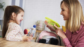 Eine Mutter und ihre Tochter sitzen an einem Tisch und packen Essen und Wasser in einen Rucksack.