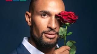 Auf dem Bild ist der aktuelle Bachelor mit einer Rose in der Hand zu sehen. Er trägt einen blauen Anzug
