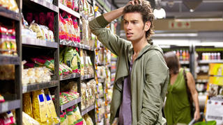 Ein Mann steht verwirrt und unschlüssig vor dem Supermarktregal.