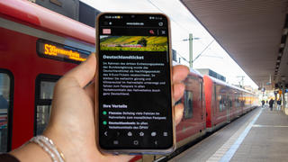 Symbolbild Deutschlandticket, 49-Euro-Ticket: Nahaufnahme eines Smartphones vor ein- und aussteigenden Fahrgästen beim Halt eines Zuges