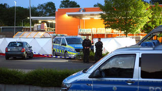 20.05.2023, Nordrhein-Westfalen, Aachen: Polizisten stehen am Tatort auf dem Parkplatz eines Baumarkts. In Aachen ist eine Frau am Samstagabend auf dem Parkplatz eines Baumarkts getötet worden. Die Polizei sperrte die Umgebung ab und setzte zur Fahndung einen Hubschrauber ein, wie ein Polizeisprecher am frühen Sonntagmorgen sagte. Schließlich wurde ein dringend Tatverdächtiger festgenommen. Die genauen Umstände und Hintergründe der Tat waren zunächst nicht bekannt. Eine Mordkommission übernahm die Ermittlungen zu dem Fall, über den zuvor die «Aachener Zeitung» berichtet hatte. Foto: Ralf Roeger/dpa - ACHTUNG: KfZ wurde aus rechtlichen Gründen gepixelt +++ dpa-Bildfunk +++