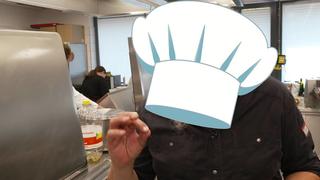 Ein Koch steht in einer Küche. Eine über seinem Kopf montierte Kochmütze verhindert, dass man ihn erkennt.