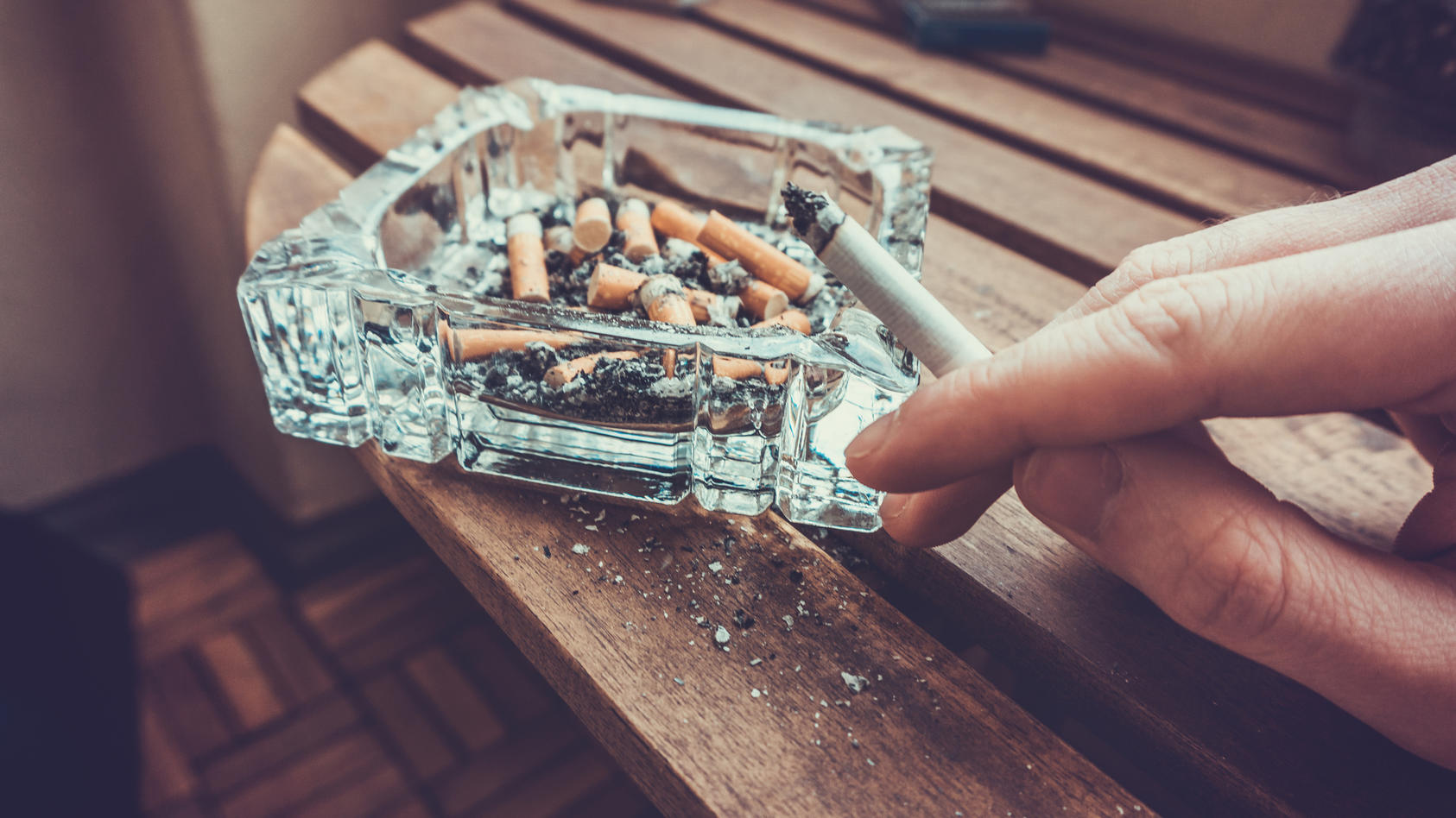 Man smoking a cigarette holding a burning filter tip in his hand alongside a glass ashtray full of ash and dead butts in a heavy smoker of addiction concept