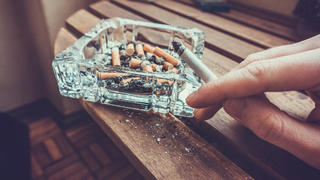 Man smoking a cigarette holding a burning filter tip in his hand alongside a glass ashtray full of ash and dead butts in a heavy smoker of addiction concept