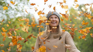 Frau in Strick-Pullover steht lachend im Blätterregen.