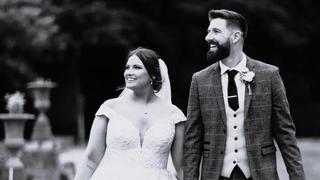 Joshua Vickers (27) im Anzug und seine Frau Laura in einem Hochzeitskleid gehen über einen Weg in einem Park.