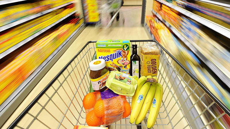 Ein Einkaufswagen mit verschiedenen Lebensmitteln wie Eier, Bananen, Orangen, Nutella, Cornflakes, Rapsöl und Toastbrot. 