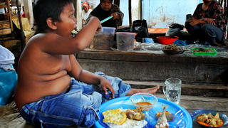 Sonderhonorar! ALDI SUGANDA hat im frühen Alter schon die ersten Laster. Rauchen und zu viel Essen! - ***EXCLUSIVE*** MUSI BANYUASIN REGENCY, INDONESIA - UNDATED: Aldi Suganda (5) is having breakfast at the market where he and his mom sell vegetable, before going to school at SDN 1 Teluk Kemang, Sungai Lilin in Musi Banyuasin, Indonesia. TODDLER Aldi Rizal stunned the world when it was revealed he had a 40-a-day smoking habit at just TWO-YEARS-OLD. The youngster was discovered in a poor village in Sumatra, Indonesia, puffing on a cigarette while riding his trike. Now five-years-old, the cheeky schoolboy has kicked the habit. But he’s picked up a new addiction – to food. His huge appetite has seen him gorge on junk food and fatty snacks - including three cans of condensed milk a day. Today he weighs nearly four stone, double what he should be for a child his age, and medics have urged his mother, Diane, 28, to put her son on a diet. 