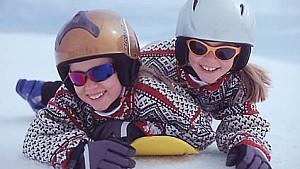 Zwei Kinder liegen auf Mini-Snowboard mit Helmen.