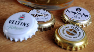 ILLUSTRATION - Kronkorken der Brauereien Krombacher, Warsteiner, Veltins und Bitburger liegen am 13.01.2014 in Köln (Nordrhein-Westfalen) neben einem Bierglas. Das Bundeskartellamt hat wegen verbotener Preisabsprachen bei Bier Bußgelder in Höhe von 106,5 Millionen Euro verhängt. Foto: Marius Becker/dpa +++(c) dpa - Bildfunk+++