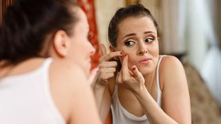 Unhappy young woman finding an acne on a cheek, sits before a mirror.