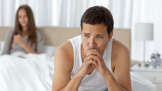 Pensive man having a headache sitting on the bed with his girlfriend on the background