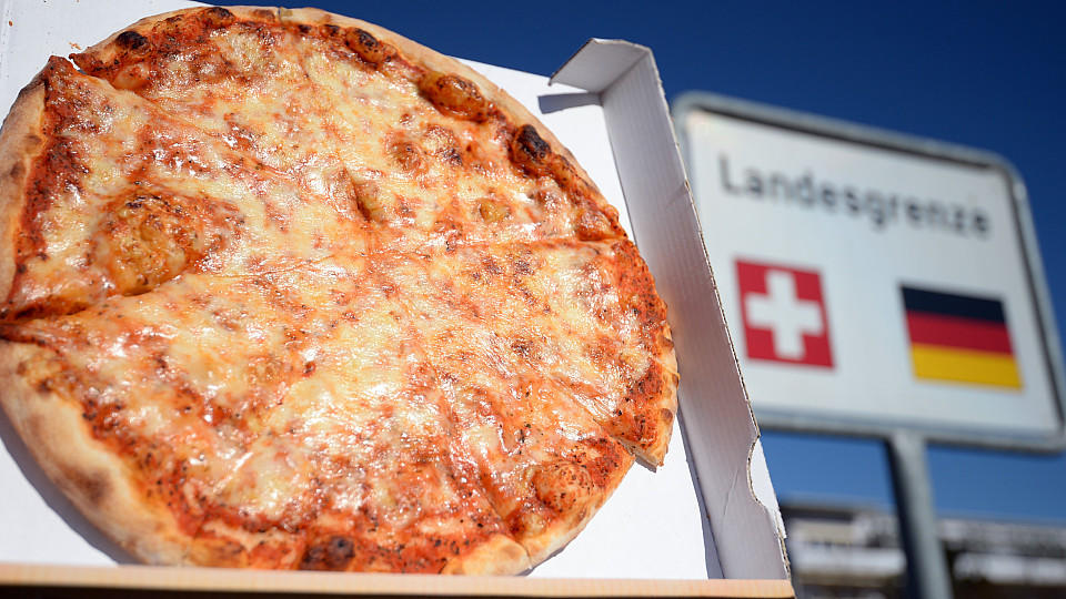 Eine Pizza vor einem Länderschild 'Deutschland-Schweiz'.