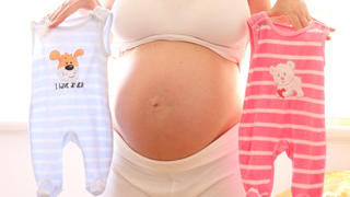 ILLUSTRATION - Eine im neunten Monat schwangere Frau hält am 20.06.2013 in Hamburg in ihrer Wohnung einen rosafarbenen und einen blauen Strampler.
