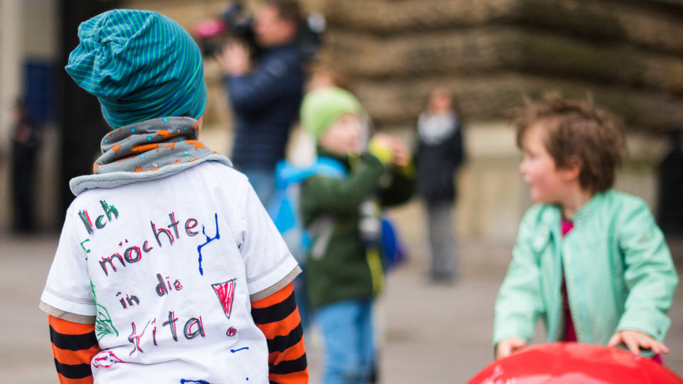 ARCHIV - Ein Kind mit einem T-Shirt mit der Aufschrift «Ich möchte in die Kita» steht am 28.05.2015 in Hamburg während einer Aktion von Eltern zum Ende des Kita-Streiks vor dem Rathaus neben Kindern, die mit Bällen spielen. Nach dem Scheitern der Sch