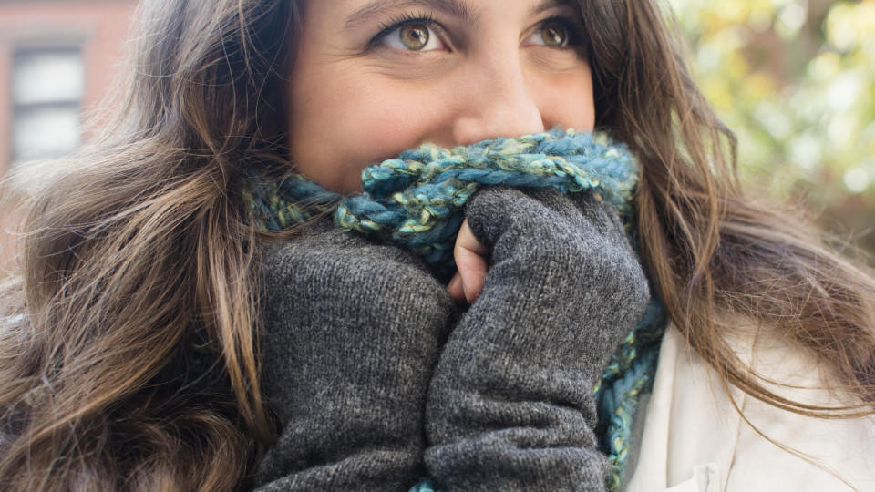 Eine junge Frau versteckt die Hälfte ihres Gesichts mit einem Schal. außerdem trägt sie Handschuhe und scheint sich gegen kalte Temperaturen zu wappnen.