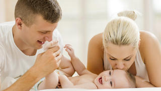 Young family enjoying together in bedroom. Father is playing with their baby boy while mother is kissing him.