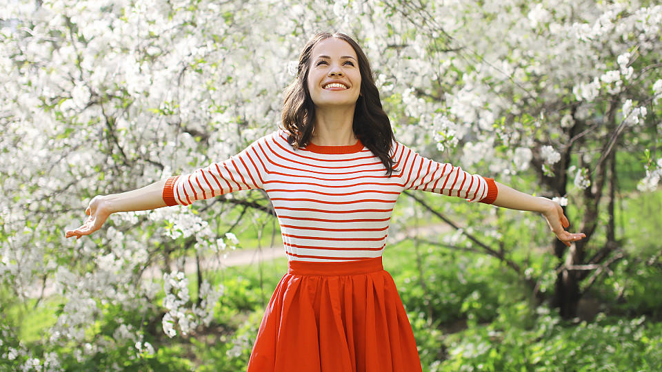 Glücklich aussehende junge Frau im rot-weiß gestreiften Shirt steht vor einem blühenden Baum