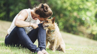 A mature Caucasian woman has relaxing fun playing with her dog in a beautiful park setting.  She lovingly talks to him with a smile on her face.  They rest in the grass, the woman arm over the dog.  Horizontal image with copy space.