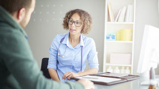 A female doctor sits at her desk and chats to a male patient about his illness . She is dressed in blouse with rolled up sleeves . She is explaining the upcoming procedure.