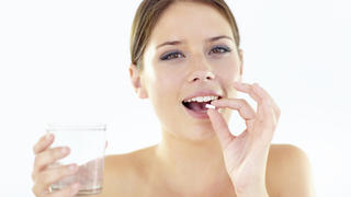 Smiling young woman isolated on white while taking a pill and holding a glass of water