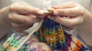Mit bunter Wolle stricken. Young woman knitting a woolen colorful scarf. She is using bamboo knitting needles, size ten. Only her hands and arms are showing on the image. She has manicured nails. Neutral colored nailpolish. She is wearing an ivory colored blouse with transparant buttons. Rolled up sleeves, bare arms are visible. The knit has pastel colors as well as brighter colors. Pink, blue, green, orange, ecru and yellow. 