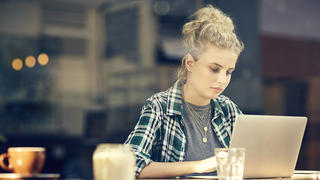 A photo of beautiful young woman using laptop at table inside cafe window. Female customer sitting in coffee shop. She is in trendy casuals.